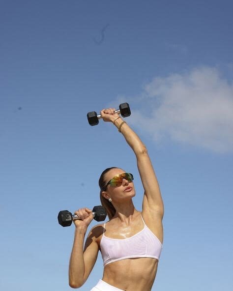 Woman lifting dumbbells overhead under a bright blue sky